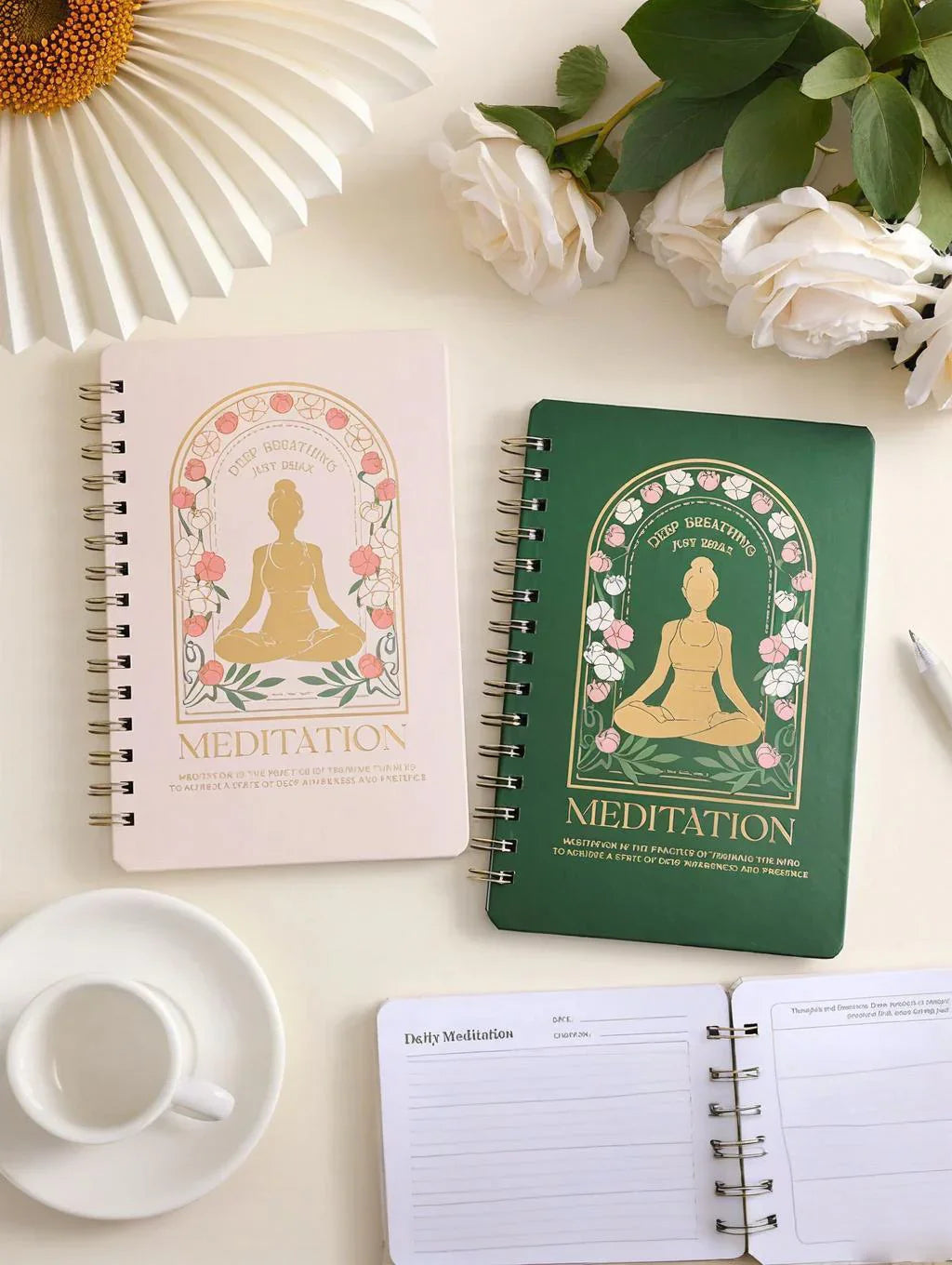 Two meditation journals with floral and yoga pose covers, white roses, a white cup and saucer, and an open journal on a white surface