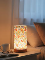 Floral-patterned lamp on a nightstand with a cup, book, and glasses in a cozy bedroom setting.