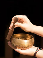 Hand holding a bronze singing bowl with a wooden striker against a dark background