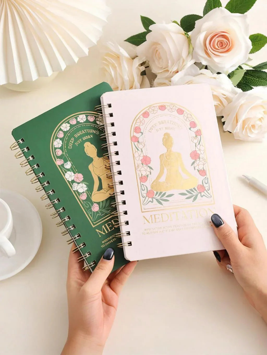 Hands holding spiral notebooks with meditation cover designs, surrounded by pale pink roses and a white cup