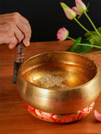 Brass singing bowl with a hand holding a mallet on a wooden surface with flowers in the background