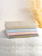 Stack of 'My Daily Self-Care' books on a textured surface with a blurred decorative background