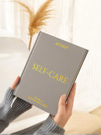 Hands holding gray self-care daily journal with gold text in cozy indoor setting