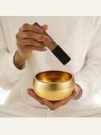 Person holding a gold singing bowl with a black and wooden striker on a white background