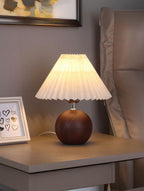 Warm-lit pleated shade lamp with a round wooden base on a bedside table (cozy night setting)