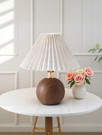 Table lamp with wooden base and pleated white shade on a round white table with a vase of flowers.