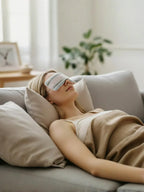Person lying on a couch with a sleep mask on, wrapped in a blanket in a cozy living room.