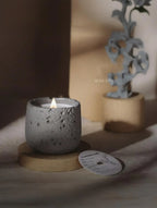 Ceramic candle holder with a lit candle on a wooden base, with a blurred plant in the background.